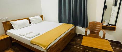 A single bed with a wooden headboard, white pillows, and a yellow blanket. There is a wooden chair and a small table beside the bed. A dark curtain hangs on the window, and a mirror is mounted on the wall.