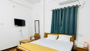 A bedroom with a wooden bed, a ceiling fan, an air conditioning unit, a TV, and a small bedside table. - Murdeshwar Comforts (Murudeshwara)
