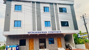 A modern building with the sign "MURDESHWAR COMFORTS" indicating a hotel. There are multiple windows and a staircase leading to the entrance. - Murdeshwar Comforts (Murudeshwara)