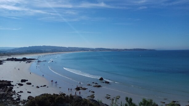 Cena de praia costeira com céu claro, costa rochosa e colinas ao longe.