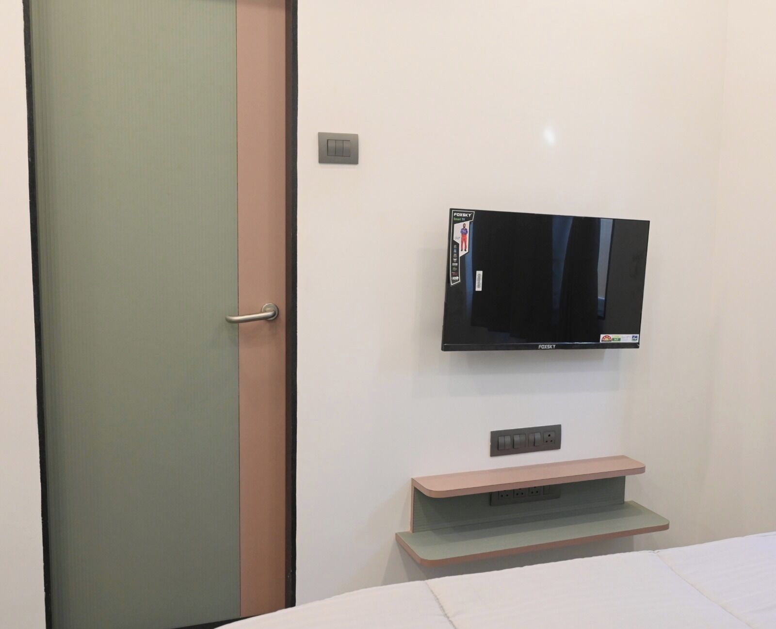 Habitación de hotel moderna con una televisión de pantalla plana montada en la pared, un mueble de televisión de madera y una puerta con un panel de vidrio esmerilado.