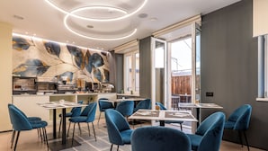 A modern dining area with blue chairs and tables, a kitchen in the background, and a large abstract mural on the wall. - Wega Hotel  (Verona)