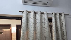 A bedroom with a bed, a window with tree-patterned curtains, an air conditioning unit, and a door with a sign. - Poonam Villas (New Delhi)