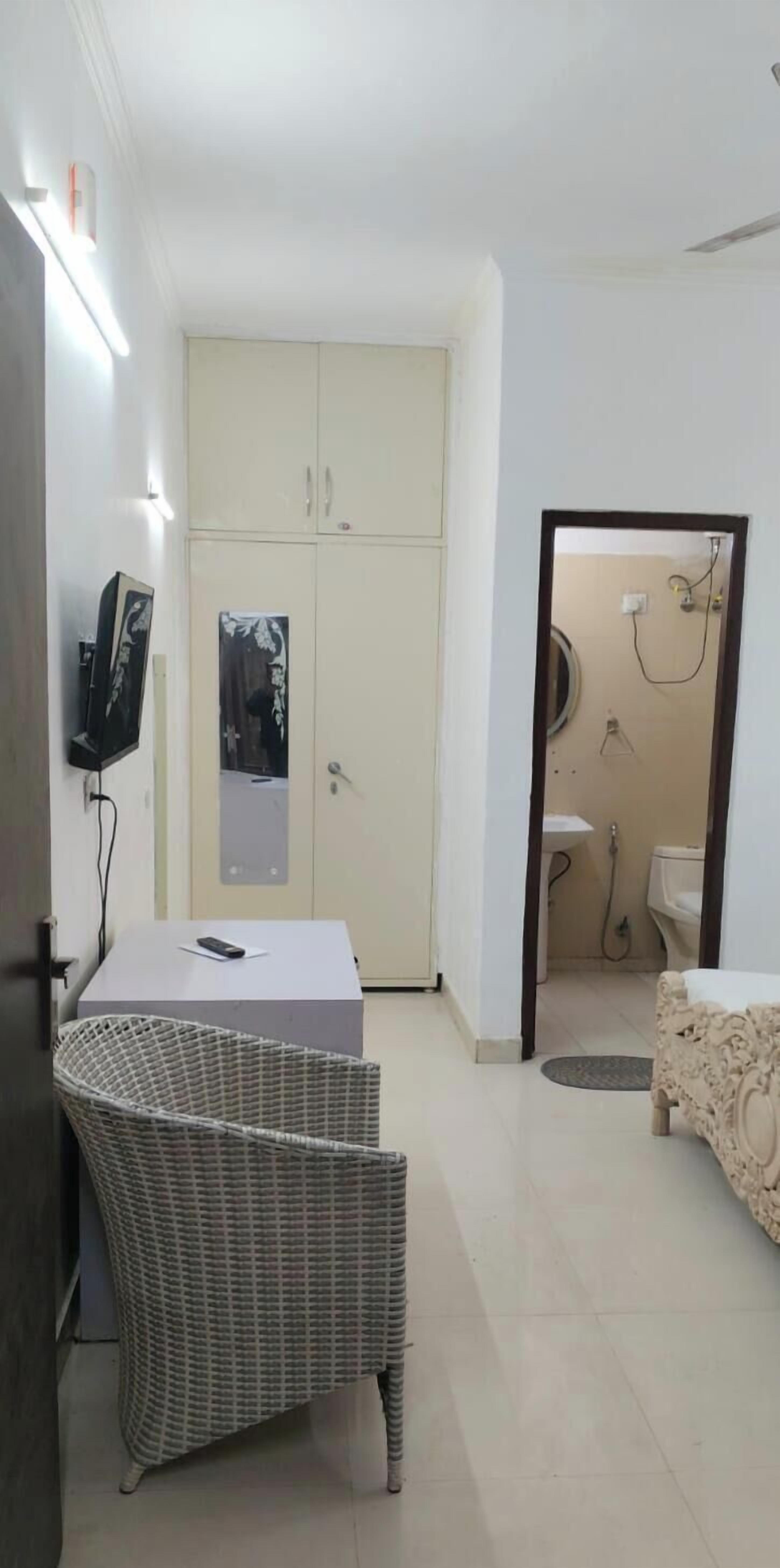 A compact room with a bed, a small table, a wicker chair, a wardrobe, and a bathroom visible through an open door.