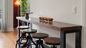 A bar area with a long wooden counter, three stools, and a small table with a lamp and bowls.
