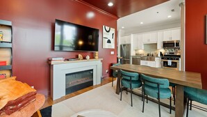 A modern living room with a fireplace, a TV mounted on the wall, and a dining area with teal chairs.