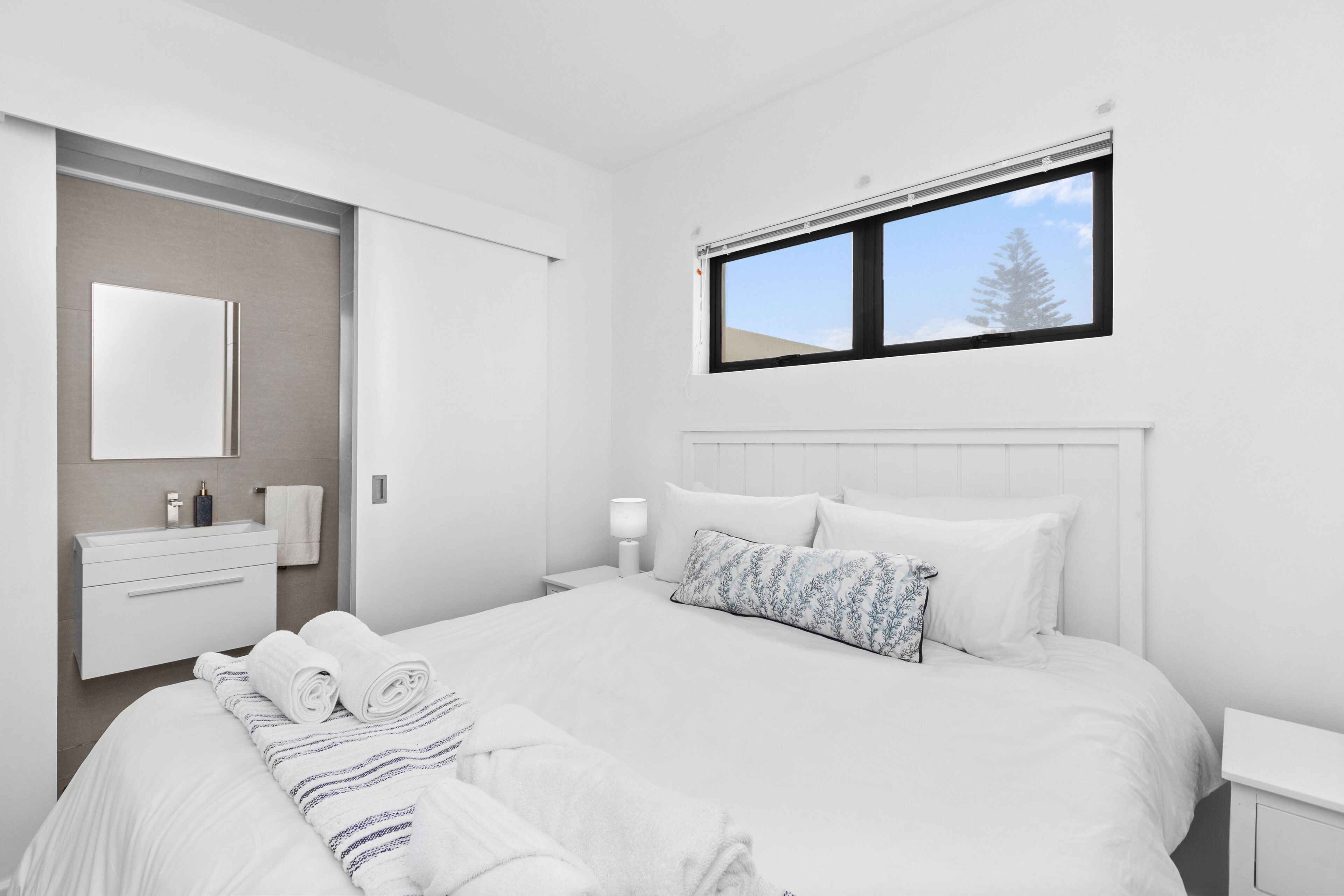 A modern bedroom with a large bed, a bedside table, a nightstand, a window, and a bathroom with a mirror and sink.