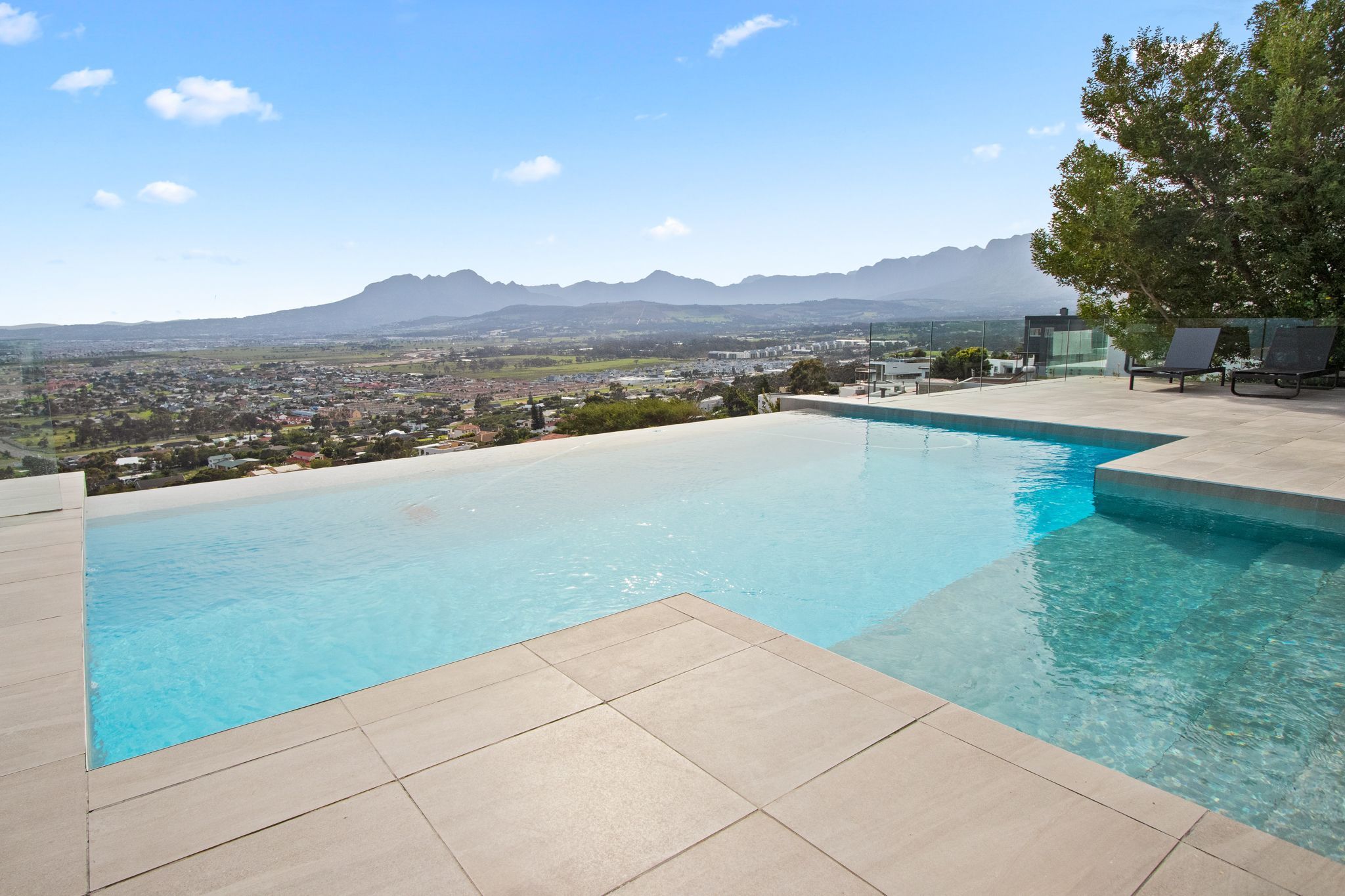 A swimming pool with clear blue water, surrounded by a tiled deck, overlooking a cityscape and mountains.