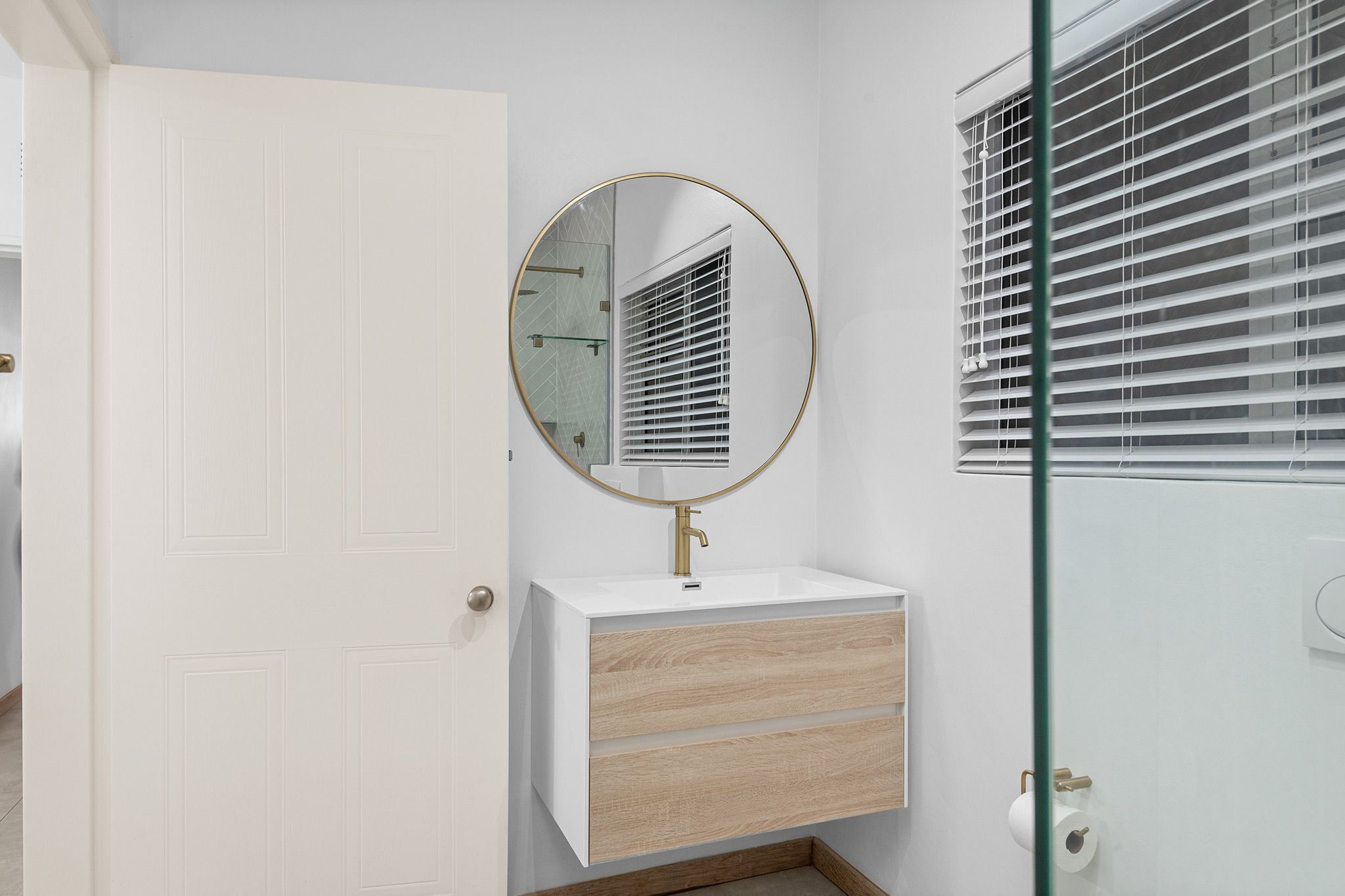 A bathroom with a white vanity, a round mirror, and a window with blinds.