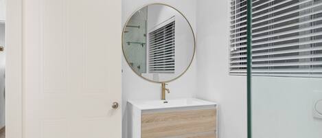 A bathroom with a white vanity, a round mirror, and a window with blinds.