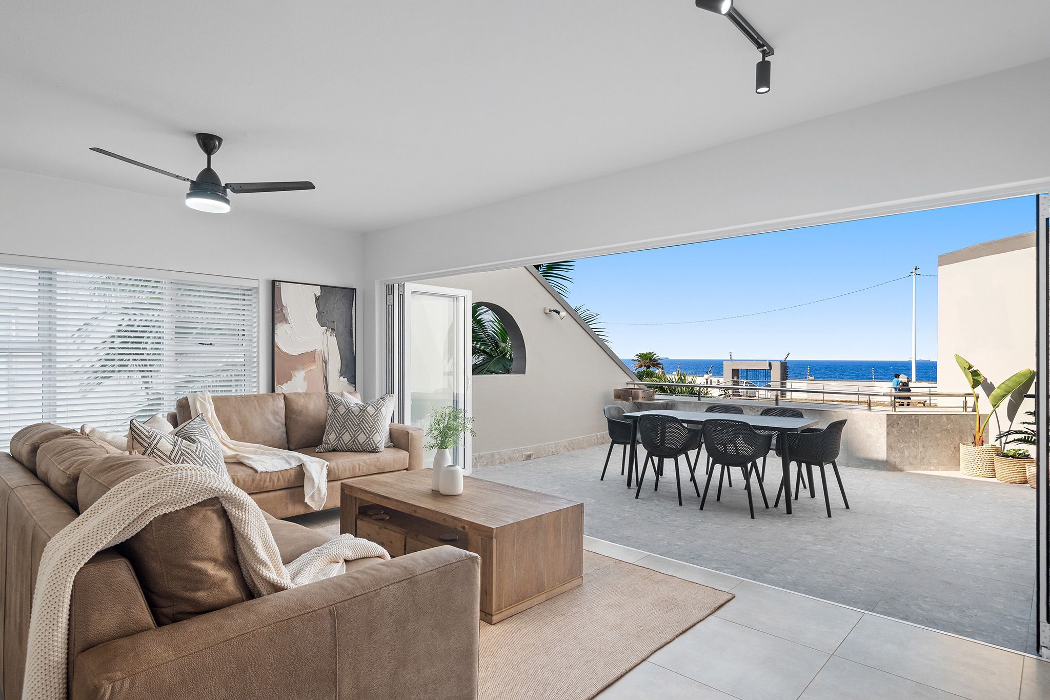 Uma sala de estar moderna com sofá, mesa de centro e área de jantar com cadeiras. Há um ventilador de teto, uma janela grande com persianas e vista para o oceano.