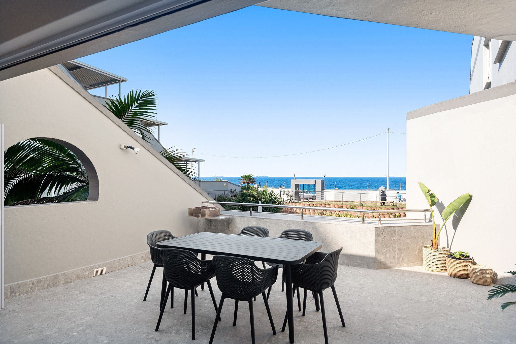 A terrace with a table and chairs, overlooking a beach and a clear sky.