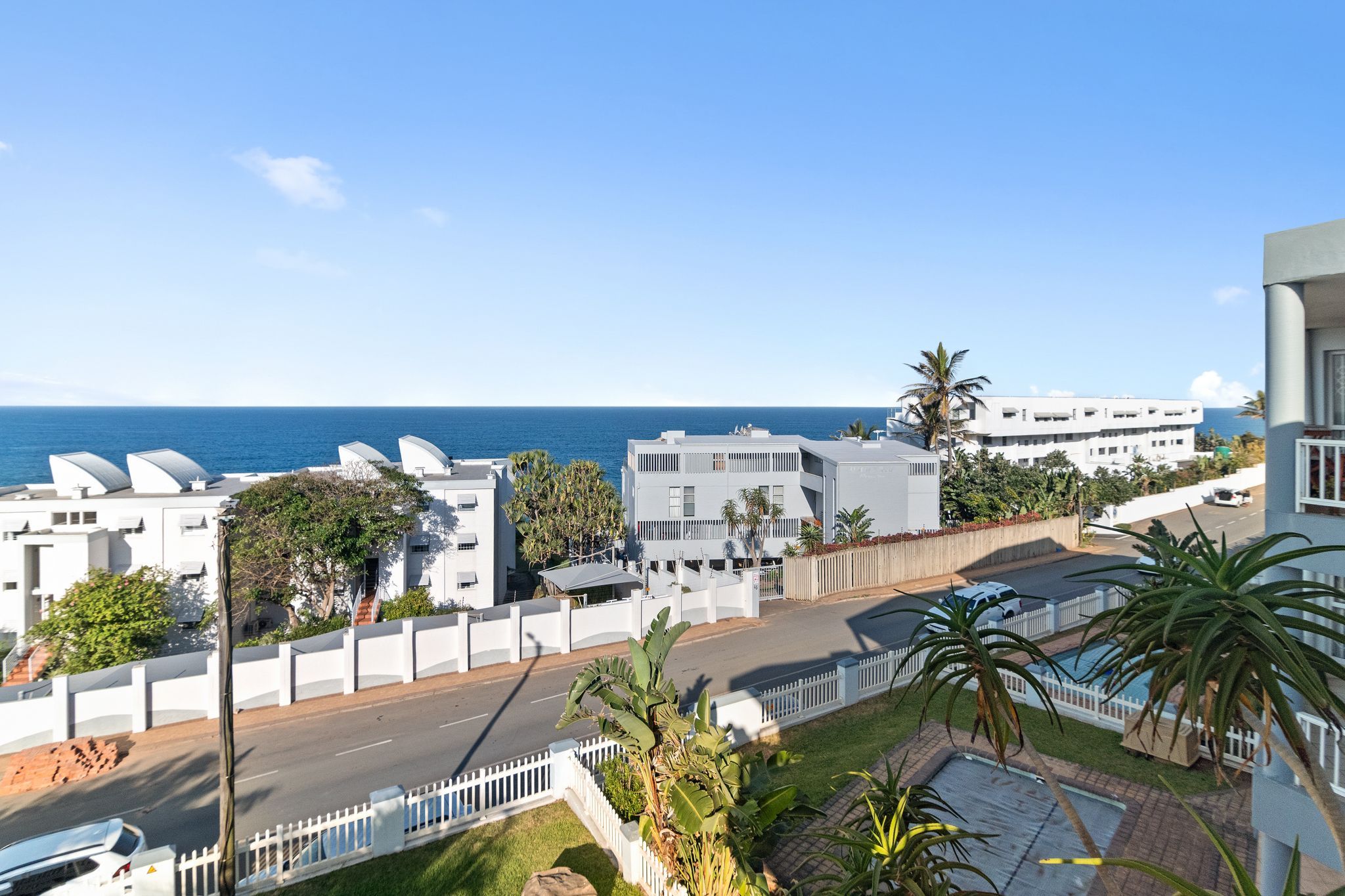 A coastal residential area with modern white buildings, a clear view of the sea, and a street with cars.