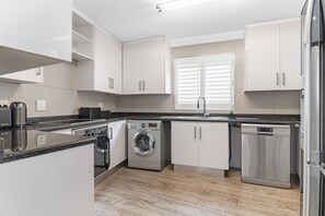 A modern kitchen with white cabinets, a built-in oven, a washing machine, and a dishwasher. - San Marino 17 (Ballito)