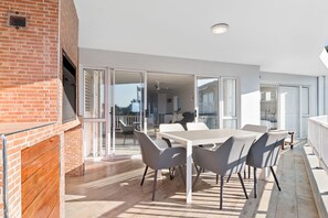 A modern outdoor patio with a white table and grey chairs, a brick wall, and a sliding glass door leading to an interior space.