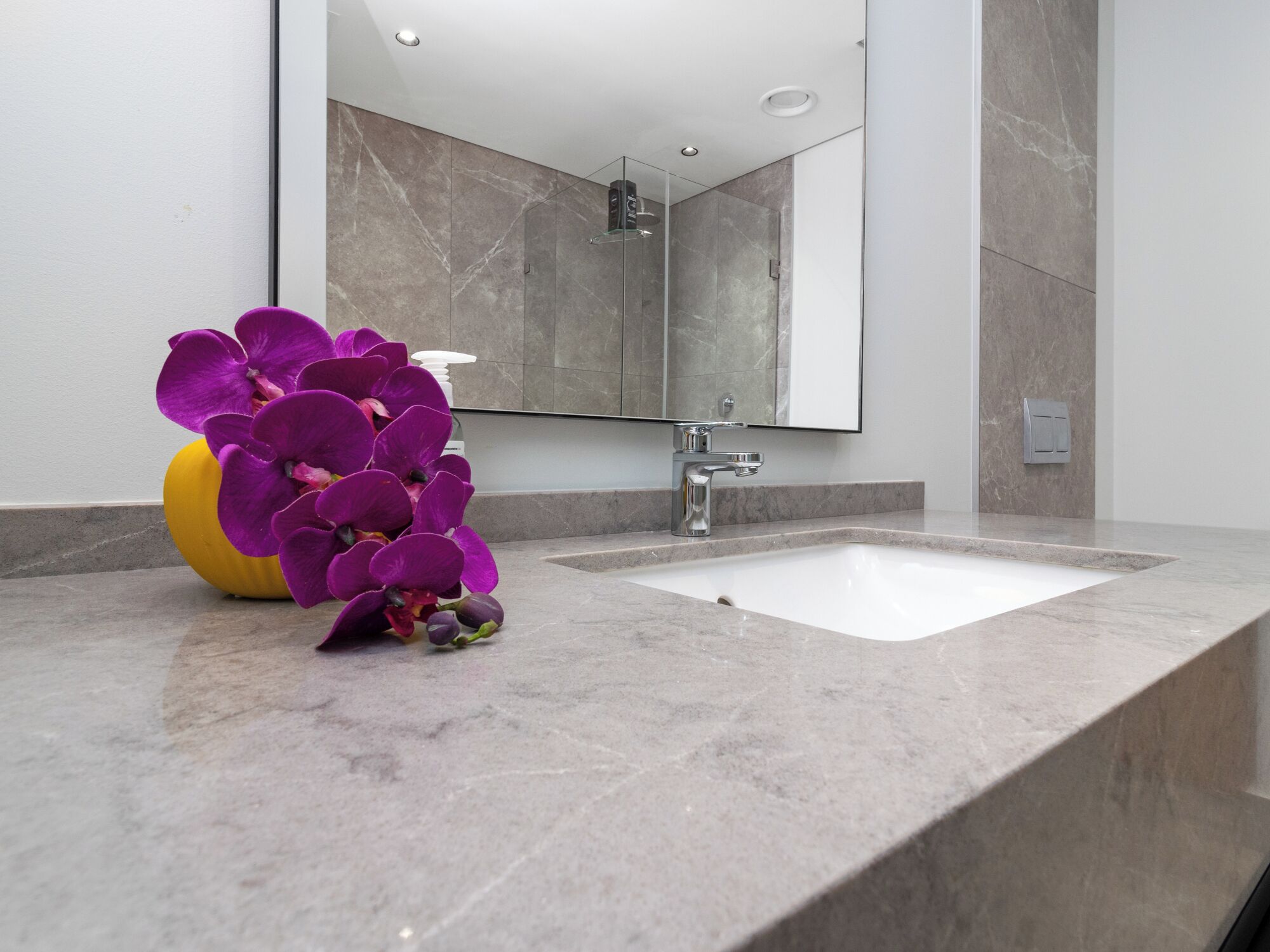 Une salle de bain avec un lavabo en marbre, un miroir et un vase rempli de fleurs violettes.