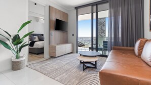 A modern living room with a sofa, a small table, a TV mounted on a wooden panel, a large window with curtains, and a potted plant. - Ellipse Waterfall 3909 (Johannesburg)