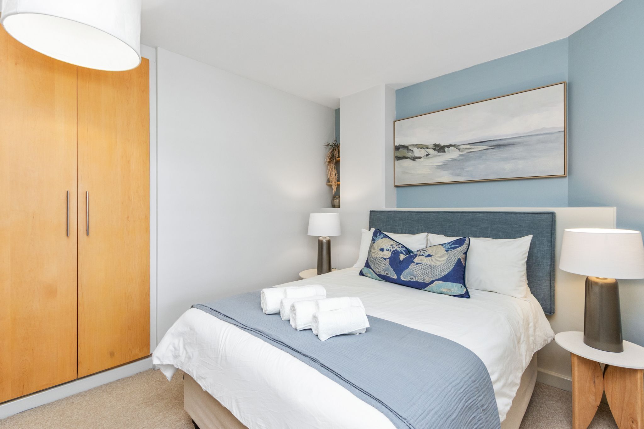 A neatly made bed with white and blue bedding, a blue headboard, and a painting above it.