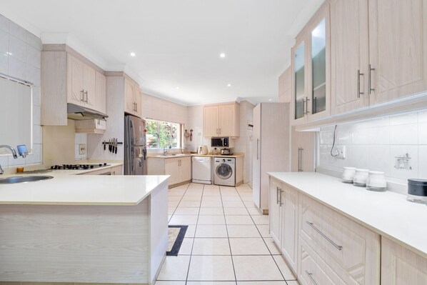 A modern kitchen with light wood cabinets, a central island, stainless steel appliances, and a view of the outdoors. - Gainsford Place 26 (uMhlanga)
