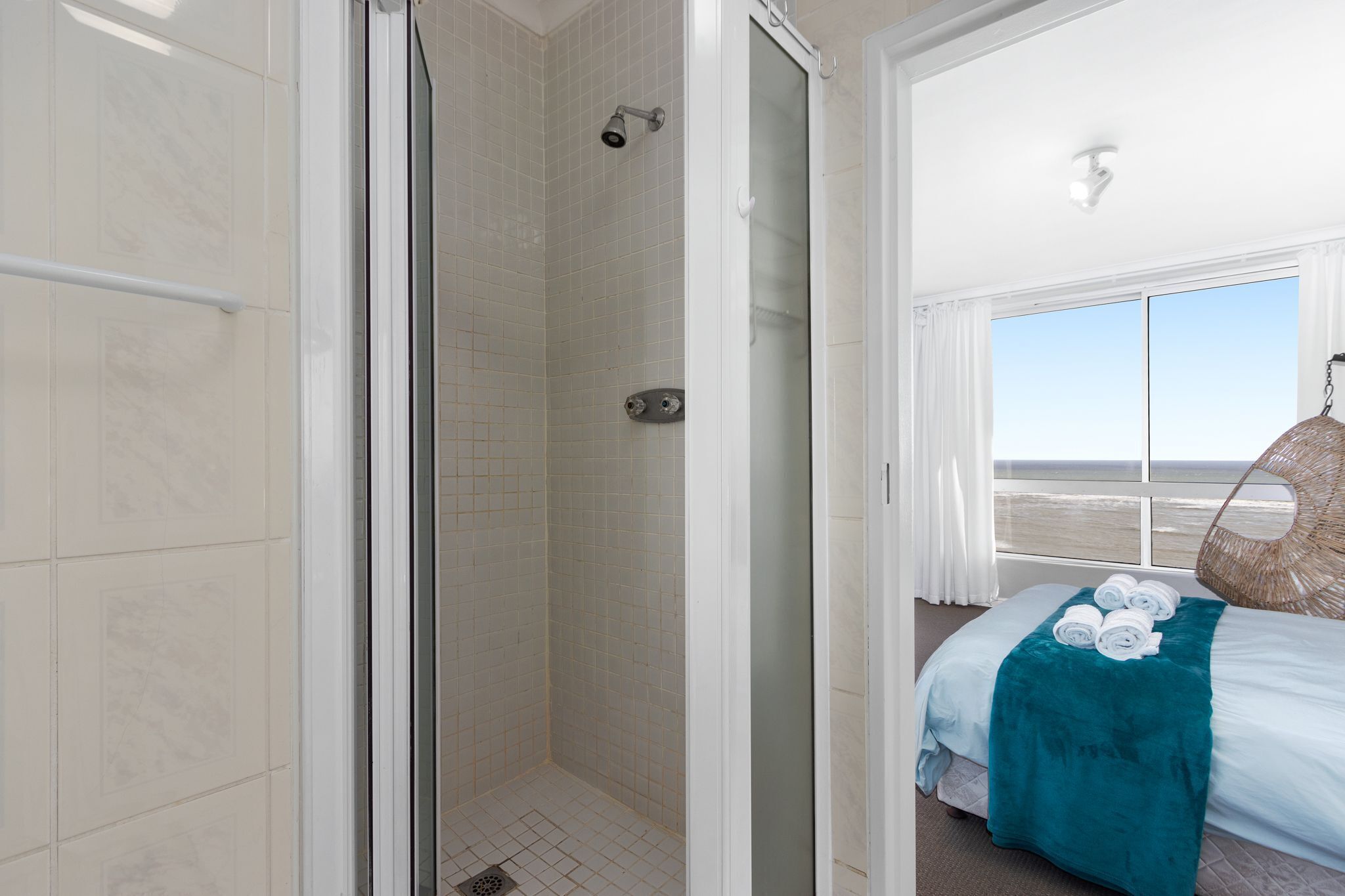 A hotel room with a glass shower enclosure, a bed with a teal blanket, and a view of the beach through a large window.