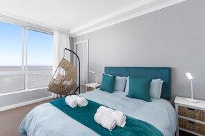 A hotel room with a large bed, teal bedding, a hanging chair, and a view of the sea. - Odeon Towers 1102 (Cape Town)