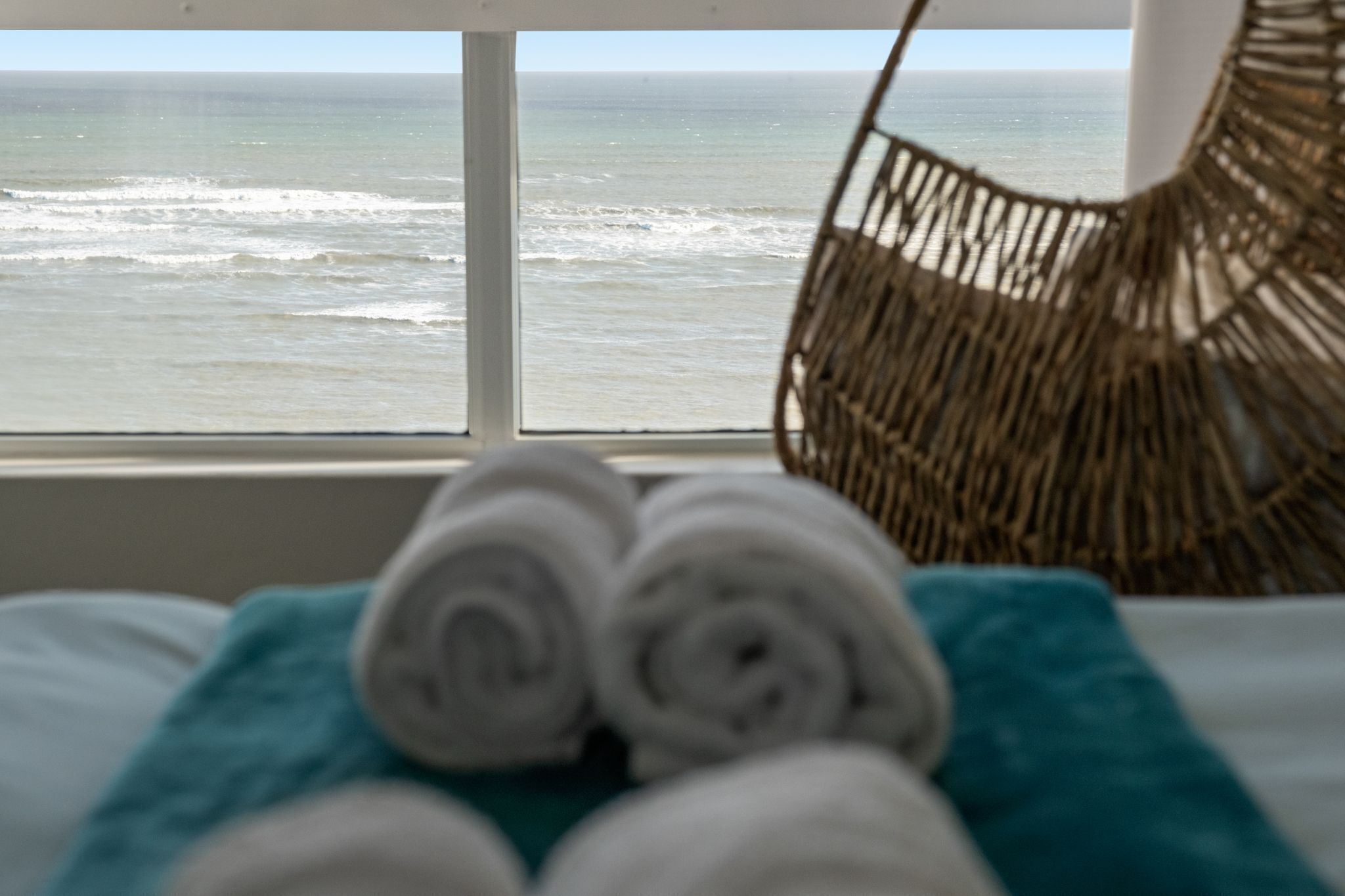 A view of the ocean through a window, with a wicker chair and rolled towels on a bed.