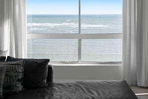 A room with a large window overlooking the ocean, a bed with a gray mattress, and white curtains. - Odeon Towers 1102 (Cape Town)