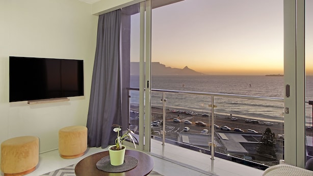 A modern hotel room with a large balcony, a flat-screen TV, and a view of the ocean and parking area.
