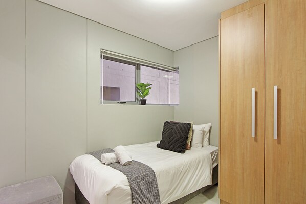 Un dormitorio con cama, una ventana con planta, un armario y un taburete pequeño.