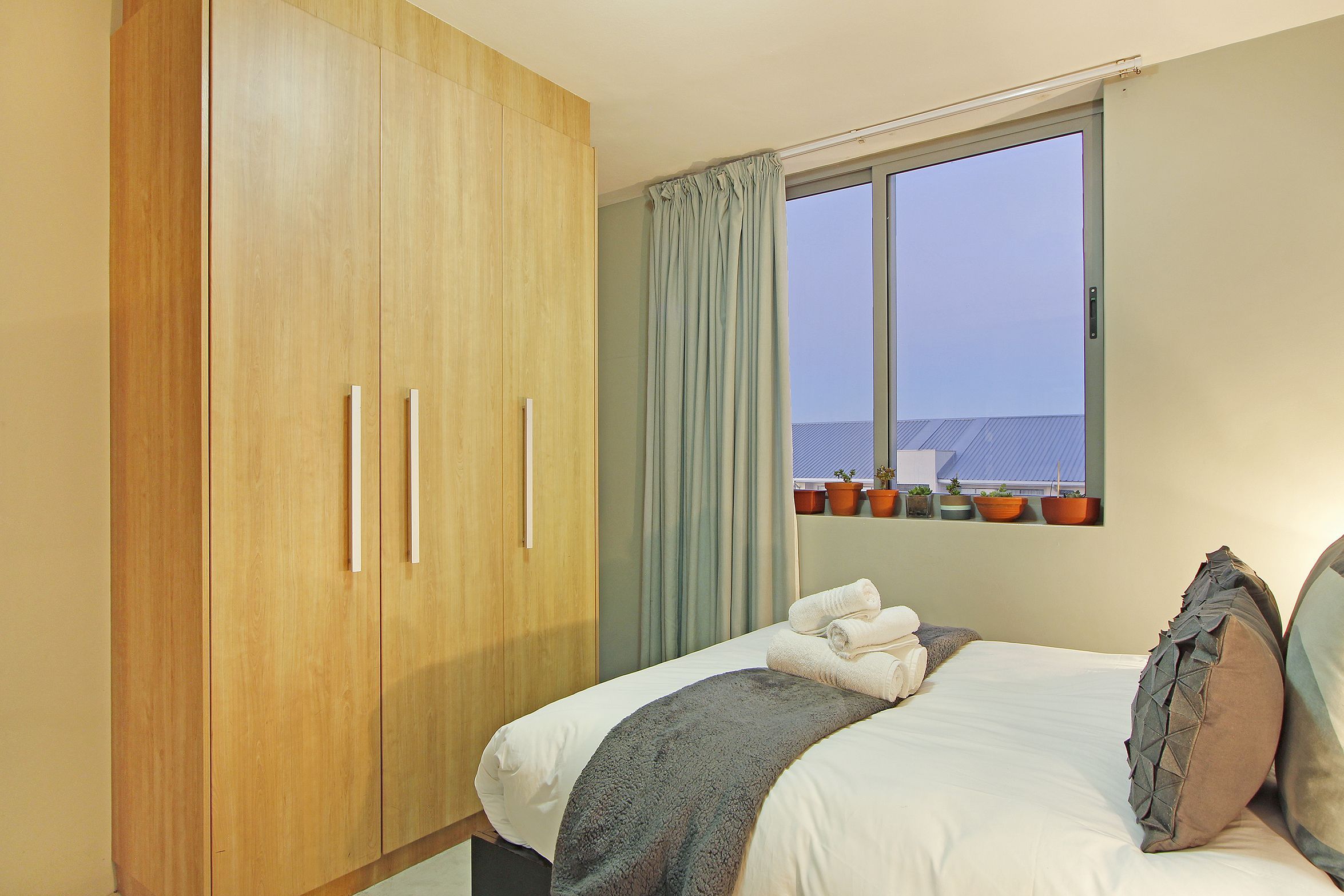 A bedroom with a large wardrobe, a bed with pillows, a blanket, and a window with curtains.