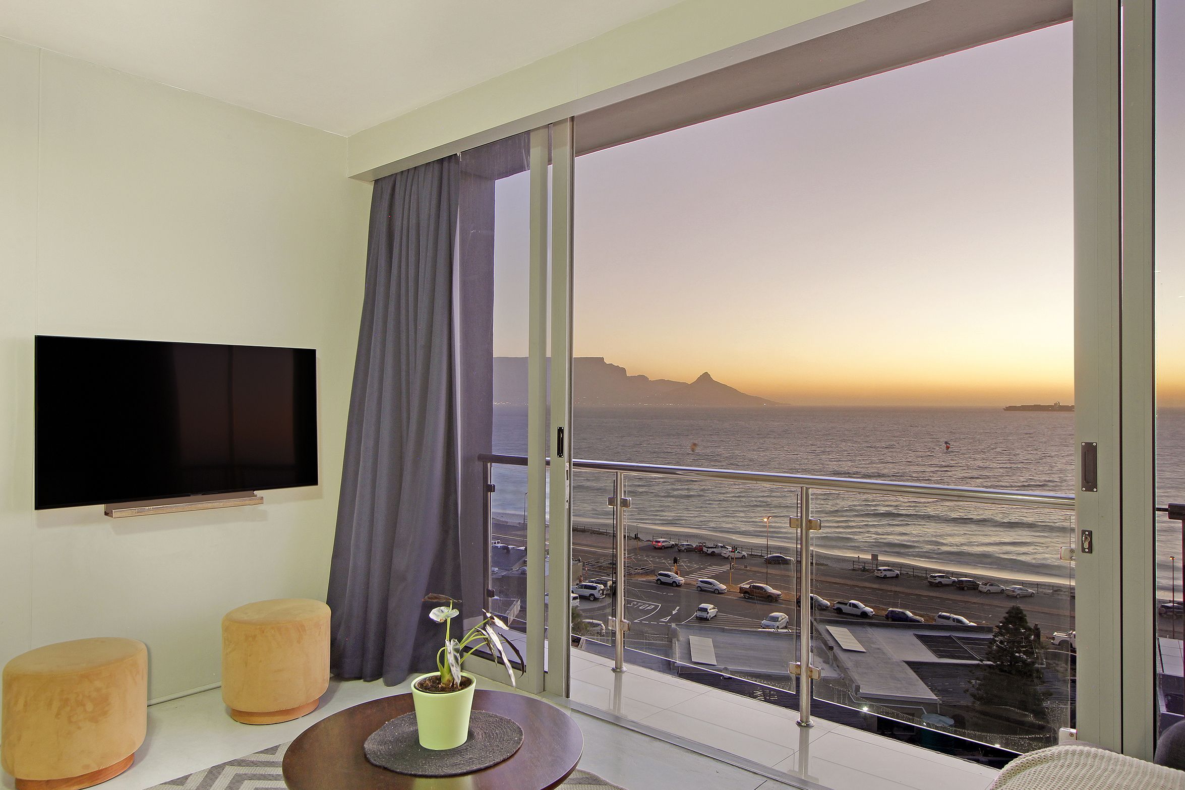 A modern hotel room with a large balcony, a flat-screen TV, and a view of the ocean and parking area.