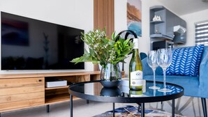 Una sala moderna con televisor, una mesa de vidrio con una botella de vino y copas, un sofá azul y una planta decorativa.