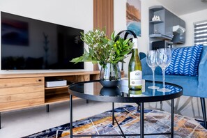 A modern living room with a TV, a round table with a bottle and glasses, a blue sofa, and a patterned rug.