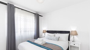 Una habitación de hotel moderna con una cama bien hecha, una mesita de noche con lámpara y un gran ventanal con cortinas.