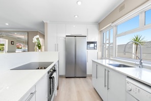 A modern kitchen with white cabinetry, a stainless steel refrigerator, a built-in oven, and a window with a view of the outside. - Ravens 9A (Cape Town)