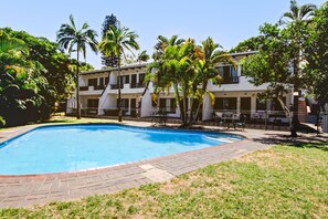 A pool area with a rectangular pool, surrounded by a brick walkway. There are palm trees and a building with multiple balconies in the background. - Villa Mia (St. Lucia)