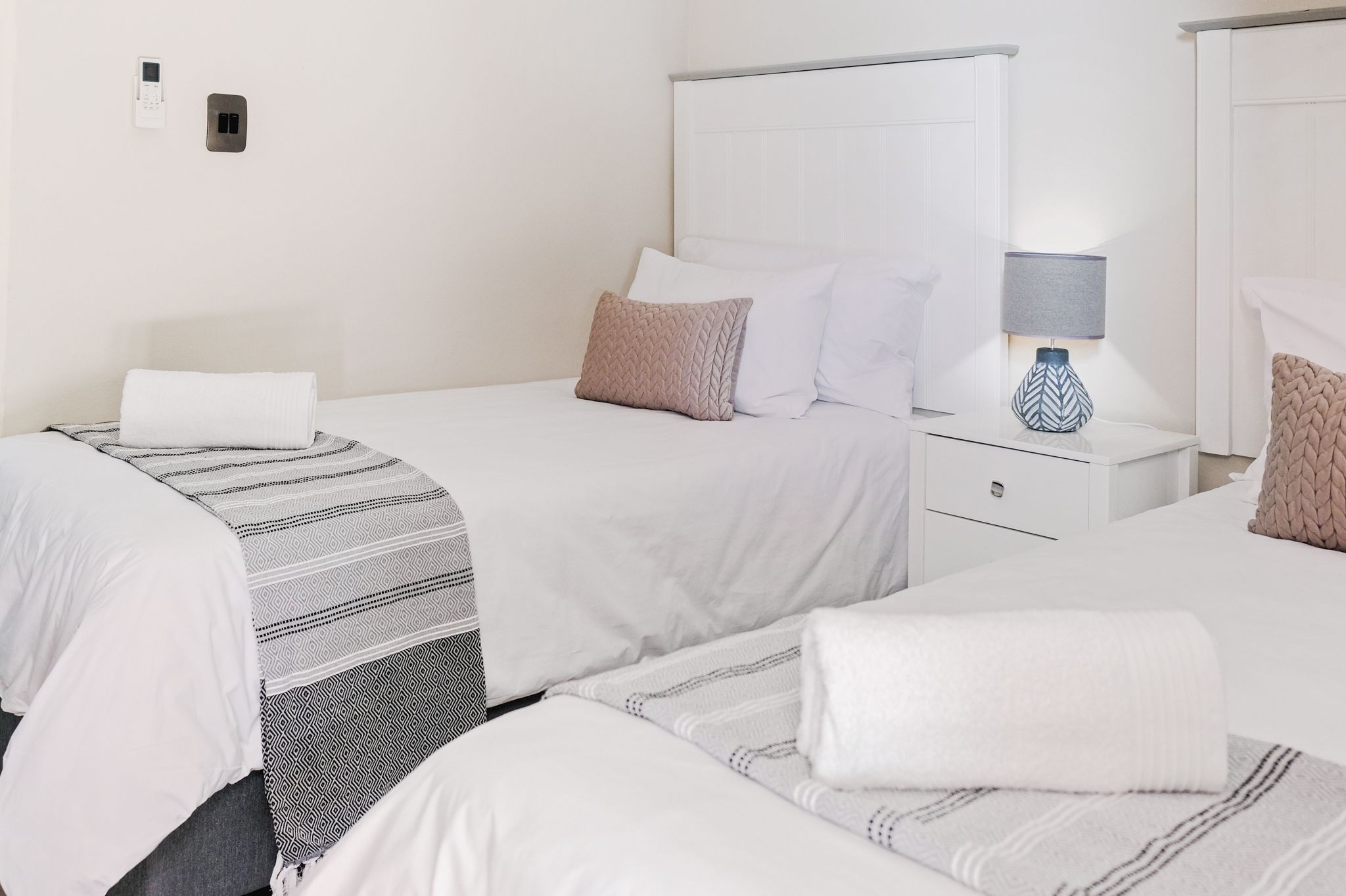 A hotel room with two beds, a bedside table with a lamp, a wall socket, and a power outlet.