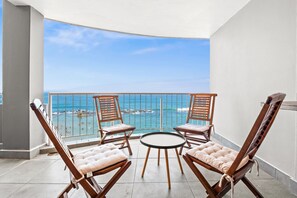 Un balcon doté de quatre fauteuils en bois et d’une petite table ronde, offrant une vue sur une plage aux eaux d’un bleu limpide et sur une côte rocheuse.