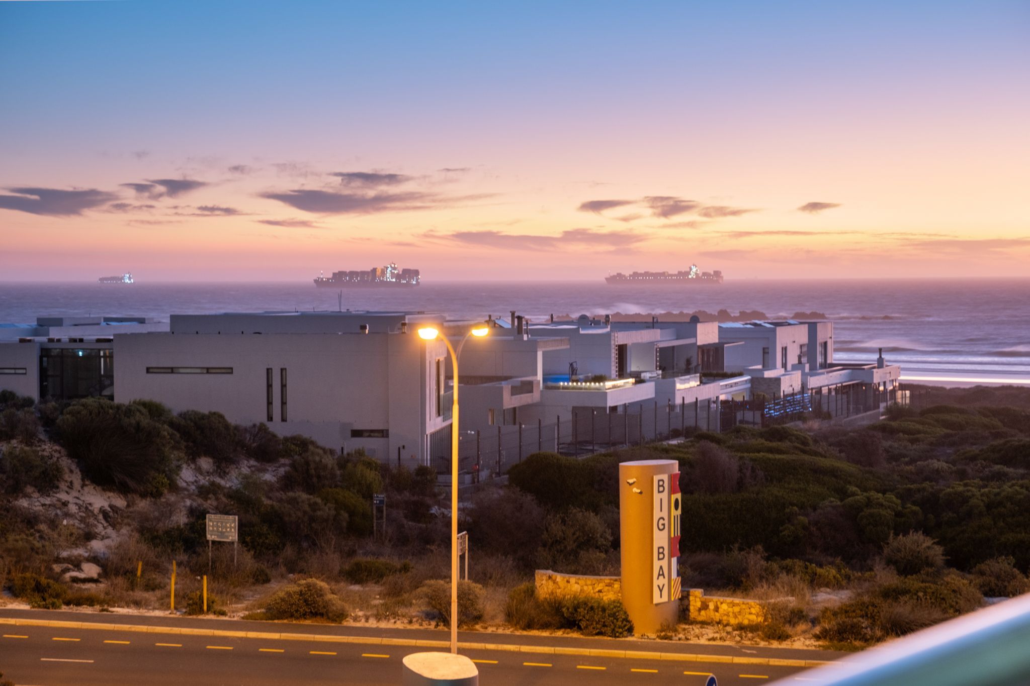 A coastal building complex with a prominent 'BIG BAY' sign, adjacent to a road with a clear view of the ocean and ships in the distance.