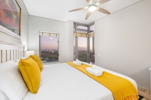 A hotel room with a large bed, a ceiling fan, and a view of the city through the windows. - Seaside Village B22 (Cape Town)