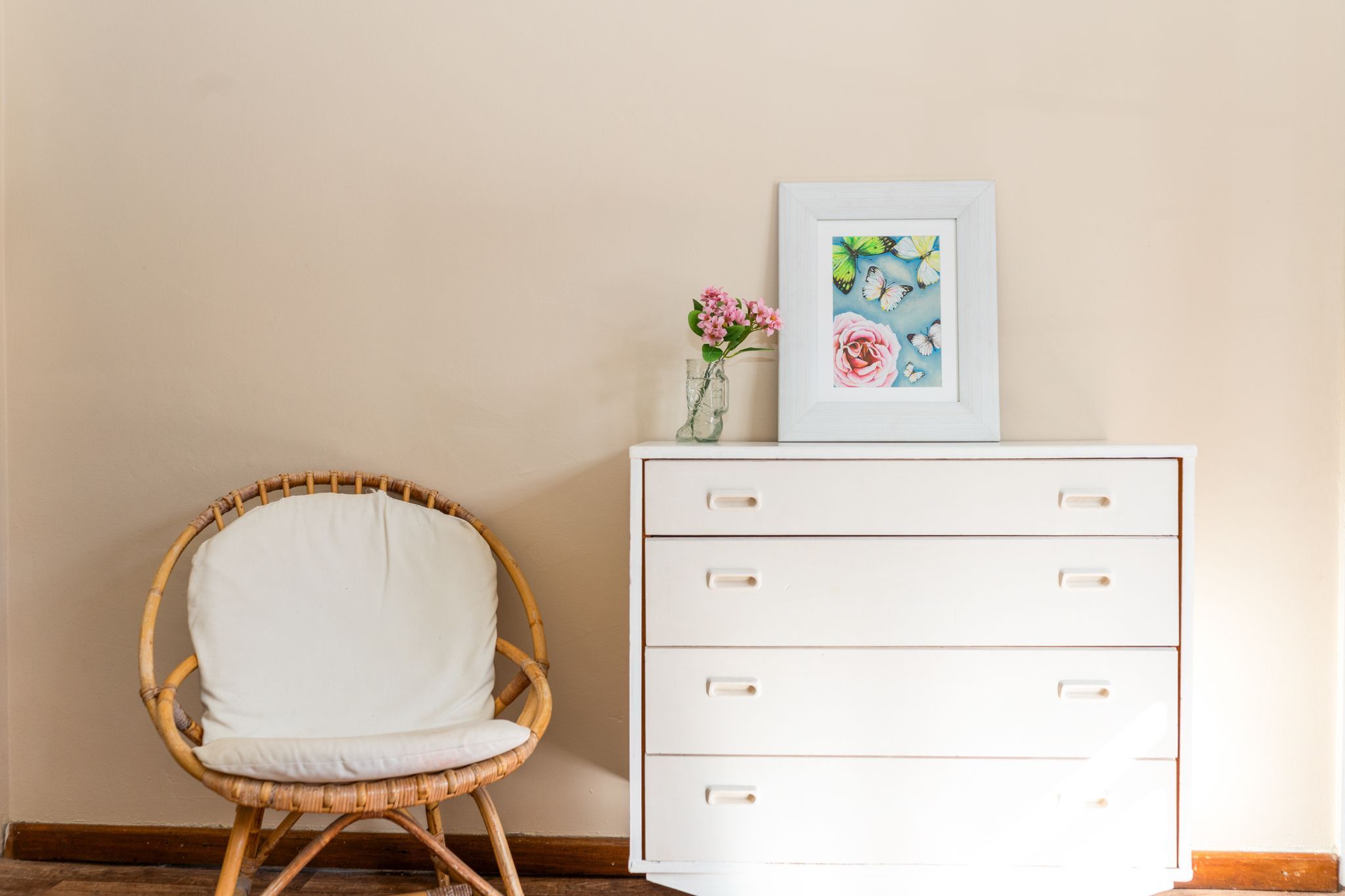 A wicker chair with a white cushion, a white dresser with drawers, and a framed picture of flowers and butterflies on the dresser.