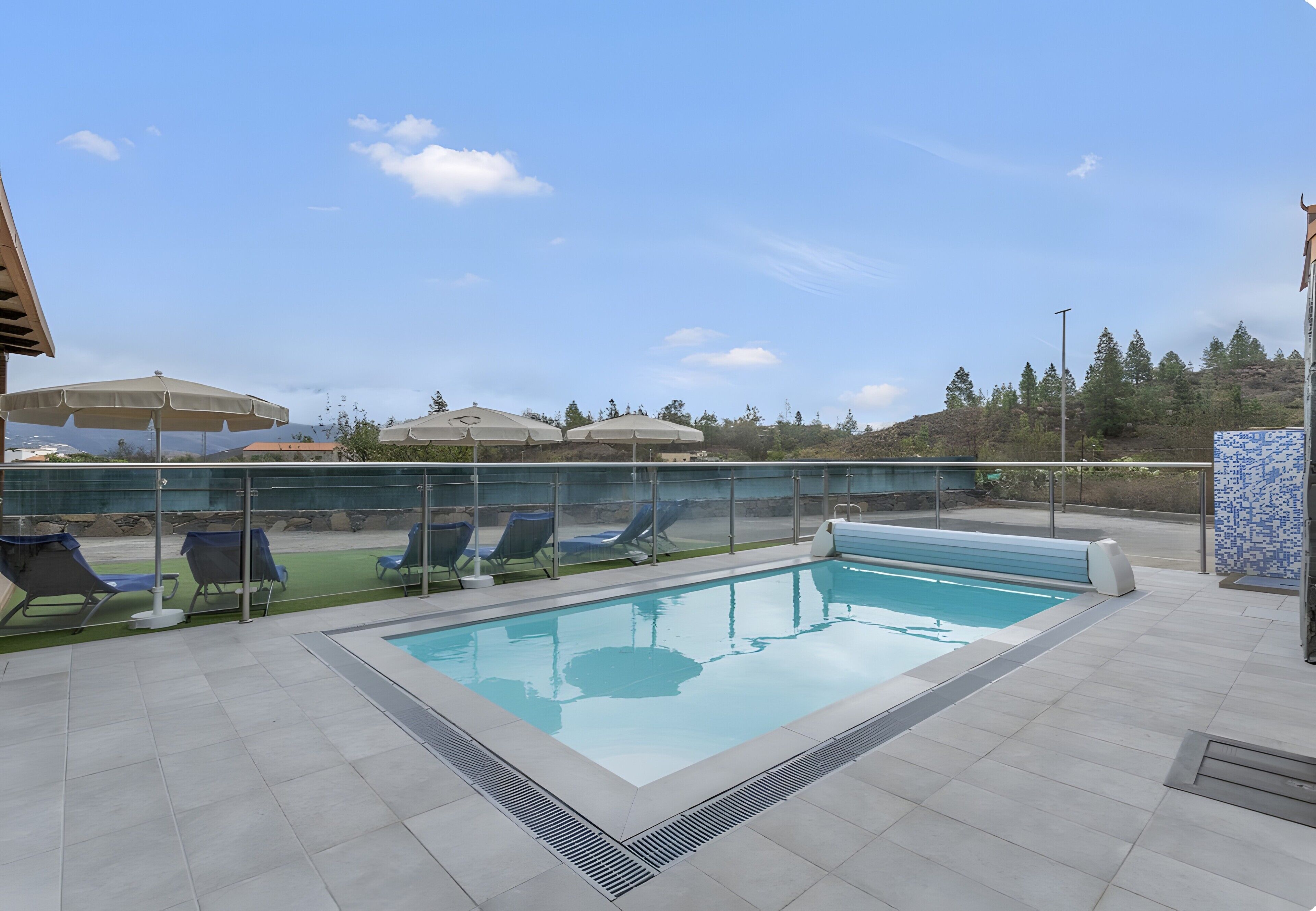 A swimming pool with lounge chairs and umbrellas, surrounded by a glass fence, and a view of trees and hills in the background.