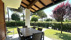 Une terrasse extérieure avec une table et des chaises en bois, entourée d’un jardin bien entretenu et de haies.