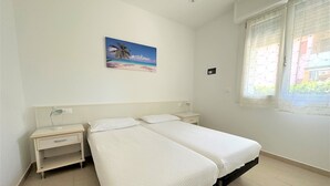 Ein Schlafzimmer mit zwei Betten, einem Nachttisch, einem Schreibtisch, einem Bild mit Strandmotiv und einem Fenster mit Vorhängen.