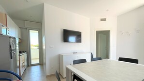 A modern kitchen with a refrigerator, stove, and cabinets. There is a dining area with a table and chairs. A television is mounted on the wall. - Great and bright flat with pool (Bibione)