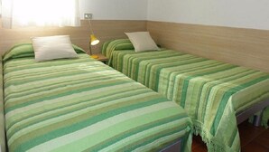 Een kamer met twee enkele bedden, elk met een groen gestreept dekbed en witte kussens. Aan één kant staat een klein nachtkastje met een gele lamp.
