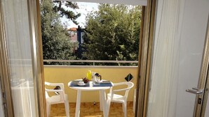 Een balkon met een tafel en stoelen, uitzicht op bomen en een gebouw, en een deur naar binnen.