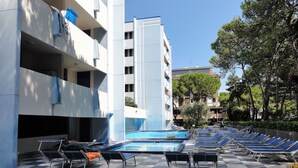 A poolside area with sun loungers and a swimming pool, surrounded by a building with balconies and trees. - Stylish Bibione flat with pool access (Bibione)