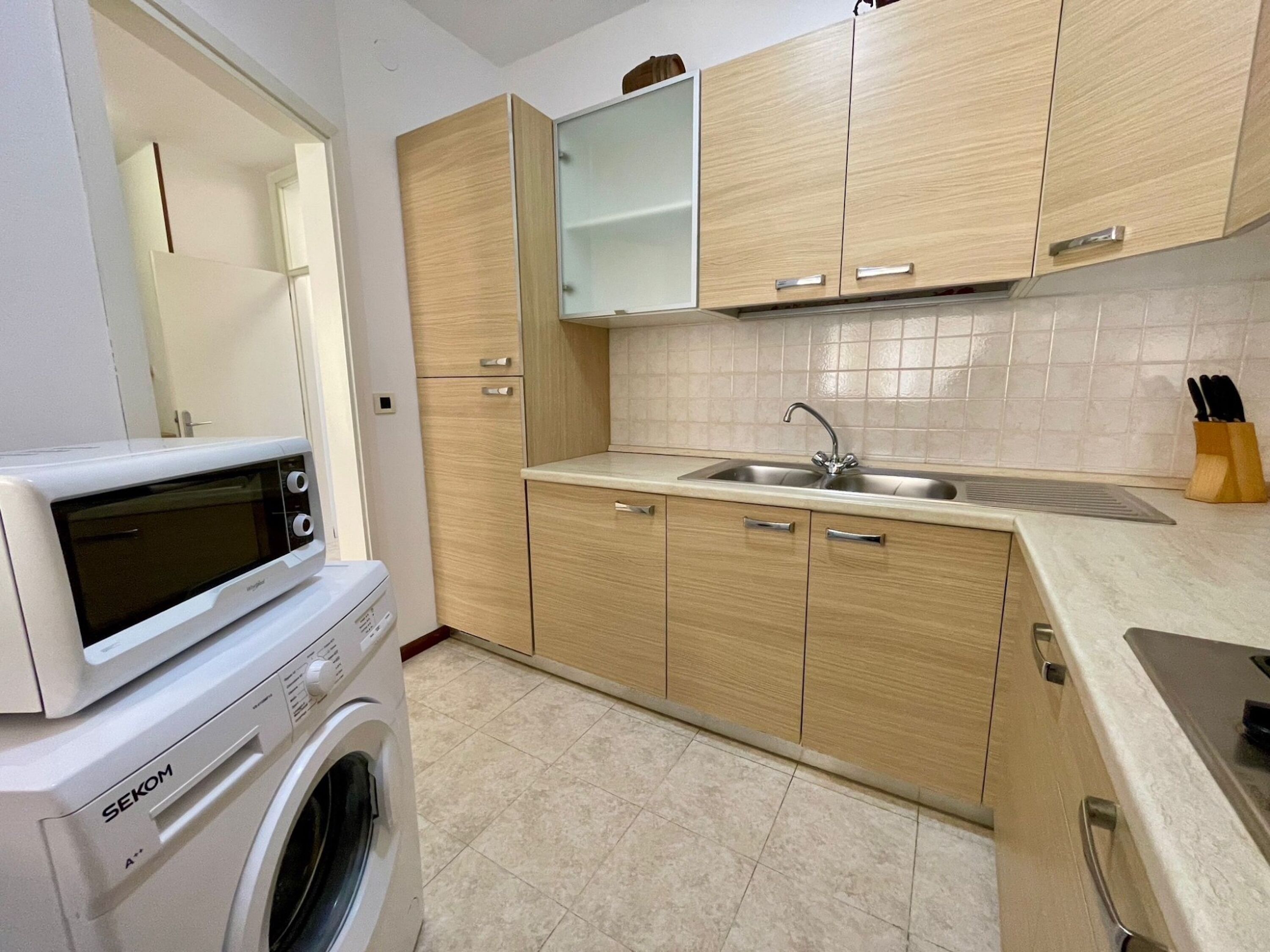 Cocina compacta con armarios de madera, microondas integrado, lavabo y lavadora.