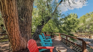 A wooden deck with Adirondack chairs and a wooden stool, surrounded by trees. - Creekside Cowa-Bungalow | Moab Studio w/ Soaking Tub (Moab)